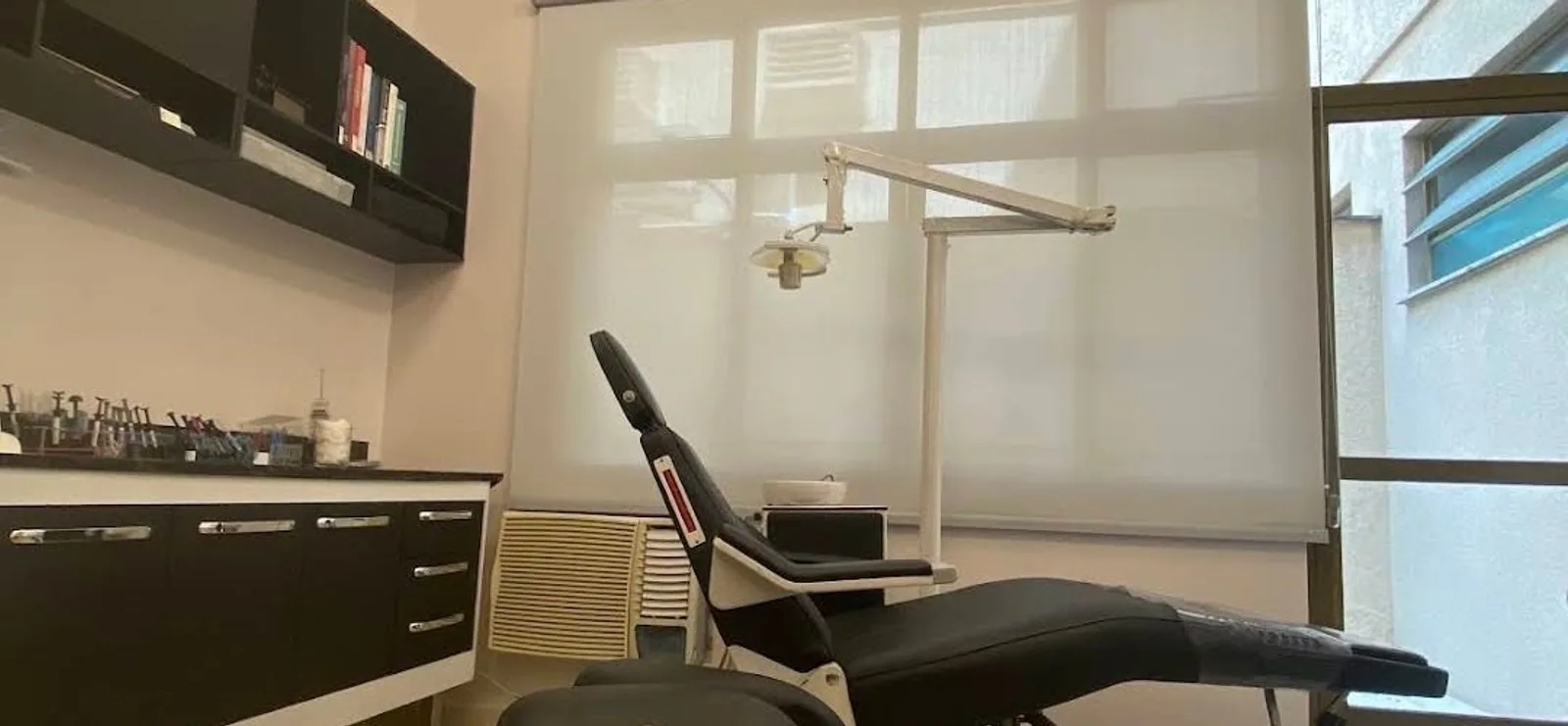 Sala de consulta da MOEBUS Odontologia em Itaipava: cadeira odontológica, armários laqueados em preto, persiana rolô, luminárias de anel quadrado em luz quente e o banco com o logo bordado.